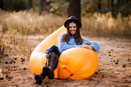 Young woman in blue sweater and black hat admires forest sunset outdoor on orange inflatable sofaの写真素材