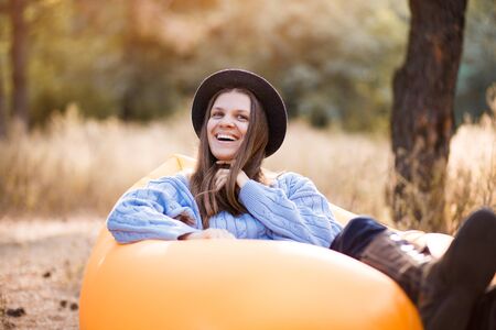 Happy autumn portrait of young smilling woman in blue sweater and black hat lying on orange inflatable sofa outdoor in forestの写真素材