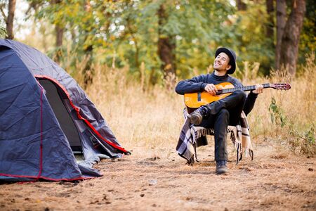 Smiling young man in black hat sitting near touristic tent and playing guitar in autumn forestの写真素材