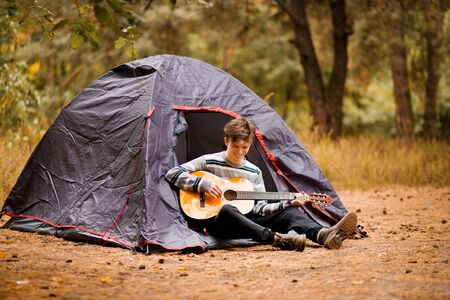 Smiling young man sitting near touristic tent and playing guitar in autumn forestの写真素材