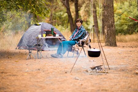 Senior woman preparing food on campfire while living in tent life in forest.の写真素材