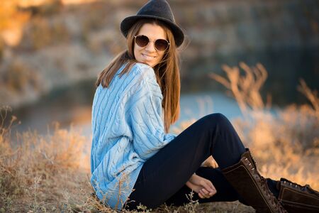 Autumn portrait of beautiful girl in sunglasses and black hat and blue sweater sitting near lakeの写真素材