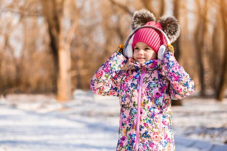Winter portrait of smiling little girl in pink hat with bubo walk in snow park and listen to music in headphonesの写真素材