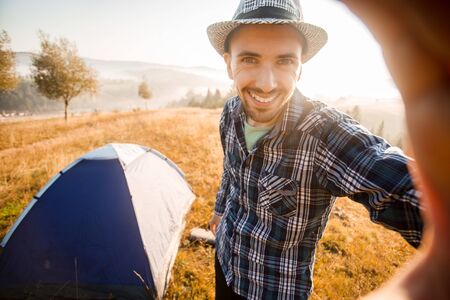 Fanny bearded man smiling and taking selfie in mountains from his smart phone. Traveler man with beard wearing hat take self portrait with camera after autumn hiking. Travel, lifestyleの写真素材
