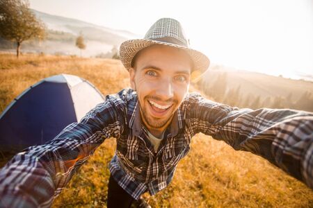 Fanny bearded man smiling and taking selfie in mountains from his smart phone. Traveler man with beard wearing hat take self portrait with camera after autumn hiking. Travel, lifestyleの写真素材