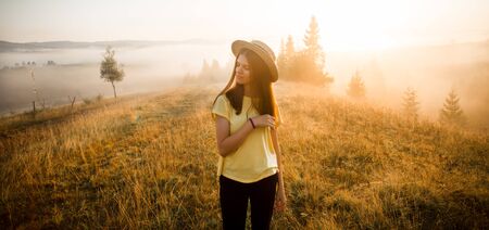 Carefree happy woman in yellow shirt and straw hat enjoying nature on grass meadow on top of mountain with sunrise. Beauty girl outdoor with sunbeams. Freedom conceptの写真素材