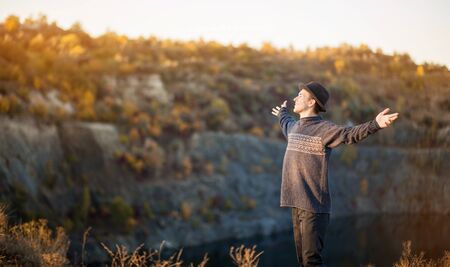Freedom concept. Free man in sweater and black hat with open arms on sunset admire nature view of autumn lake. success. Travel. Good life.の写真素材