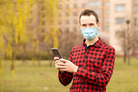 Caucasian man wearing shirt and protective mask against virus standing outdoor using smart phone mobile to send messages browsing internet video callの写真素材