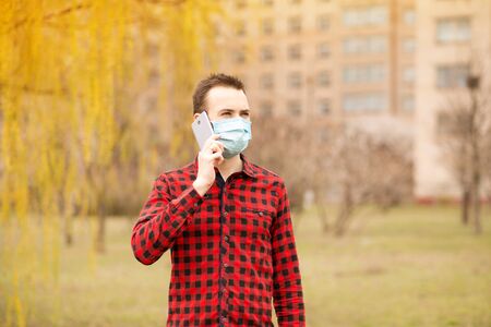 European young man in medical mask on his face calling by phone. Man in protective mask is protected from virus outdoor in streetの写真素材