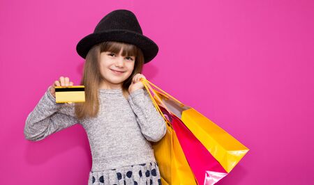 Little beautiful child girl in black hat holding shopping bags and credit card isolated on pink background. Online shopping without leaving homeの写真素材