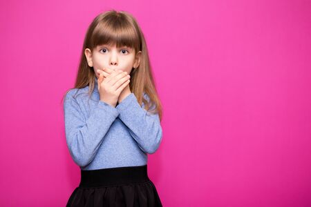Cute little girl covering her mouth showing intense expression of fear isolated on pink backgroundの写真素材