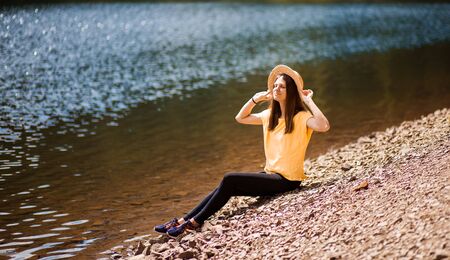 Freedom. Happy young woman sitting on beach of forest lake with arms raised, outdoor.の写真素材
