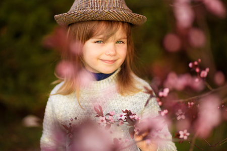 Spring portrait, adorable little girl in hat walk in blossom tree garden on sunset