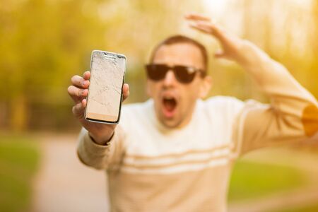 Handsome young man hold broken smartphone screen stressed with hand on head, shocked with shame and surprise face, angry and frustrated. Fear and upset for mistake.の写真素材