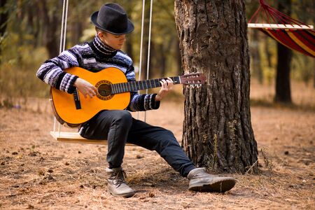 young handsome attractive man in sweater playing guitar in forest sitting on swing with hammock on background. Casual man, life style.の写真素材