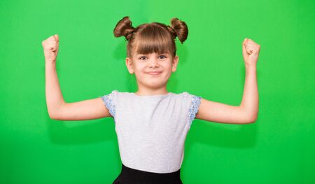 Positive little girl with funny pigtails shows her power biceps with arms raised victory. Winner school girl celebrates success, energy, yes isolated on green backgroundの写真素材