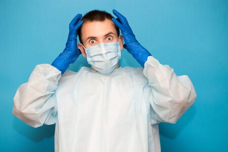 Portrait of scared man in protective suitt, medical mask and gloves against viruses and infections isolated on blue background.の写真素材