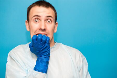 Portrait of scared man in protective suitt, medical mask and gloves against viruses and infections cover face by hand isolated on blue background.の写真素材