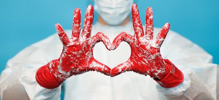 Doctor in medical suit protective mask and gloves, show heart symbol with hand in soap foam isolated on blue background. Hands hygiene, virus spreading pandemic prevention. Safe life conceptの写真素材