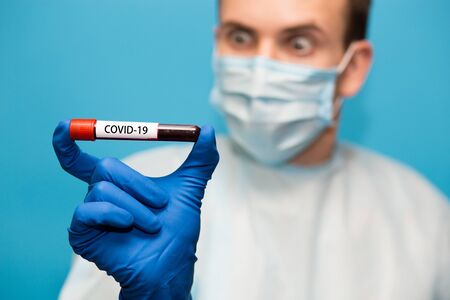 Doctor in protective mask and gloves isolated on blue background holds test tube containing blood sample that has tested for coronavirus. Laboratory testing patients blood for presence of coronavirus COVID-19.の写真素材