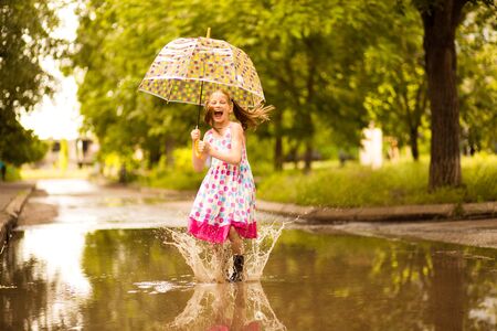Happy funny kid girl with umbrella jumping on puddles in rubber boots and in polka dot dress and laughingの写真素材