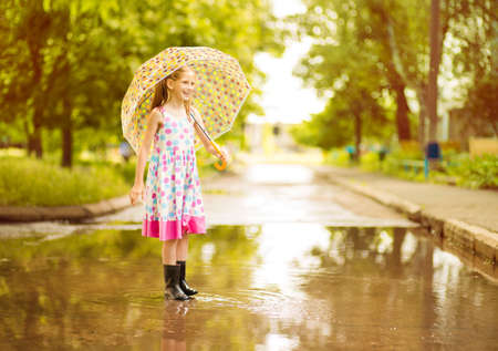 Happy funny kid girl with umbrella jumping on puddles in rubber boots and in polka dot dress and laughingの写真素材