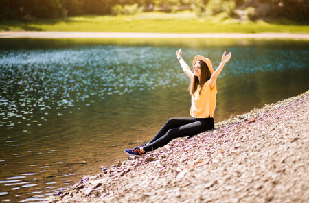 Freedom. Happy young woman sitting on beach of forest lake with arms raised, outdoor.