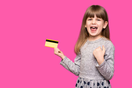 Smiling Pretty child girl with hands up in winner pose holding credit card while rejoices isolated over purple background. Mock up copy space. Doing winner gestureの写真素材