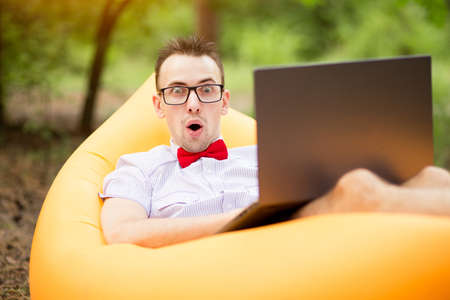 Outdoor portrait of man with surprised face in shirt, red bow tie and glasses sitting on inflatable sofa with laptop in summer park. Concept freelancing, teleworkの写真素材