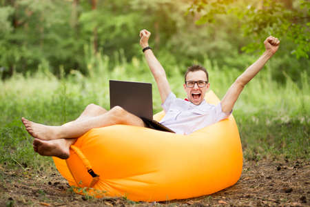 Barefoot man with hands up in winner pose and happy screaming, in shirt, red bow tie and glasses lying on inflatable sofa with laptop in summer park.の写真素材