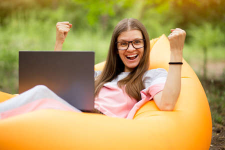 Woman with hands up in winner pose and happy screaming, in glasses lying on inflatable sofa with laptop in summer park. Teleworkの写真素材