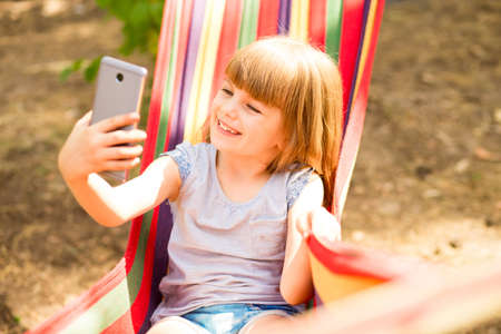 Cute little girl walking outdoors and rest on hammock in summer forest taking selfie or make video call with cell phone.の写真素材