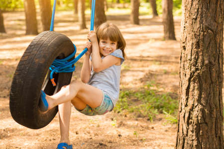 Happy active child girl playing on swing wheel in forest on sunny summer day. Summer outdoors activity for kids. Preschool child having fun and swinging on a tireの写真素材