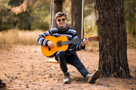 young handsome attractive man in sweater and black hat playing guitar in forest sitting on swing. Casual man, life style.の写真素材