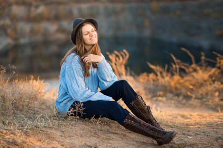 Autumn portrait of beautiful girl in black hat and blue sweater sitting near lakeの写真素材