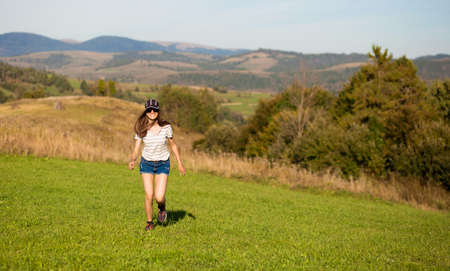 Carefree happy woman in sunglasses, cap and jeans shorts walk on green grass meadow on top of mountain enjoying nature. Freedom. Enjoyment. Daydreamingの写真素材