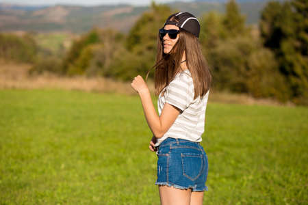 Carefree happy woman in sunglasses, cap and jeans shorts walk on green grass meadow on top of mountain enjoying nature. Freedom. Enjoyment. Daydreamingの写真素材
