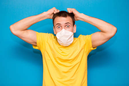 Portrait of young embarrassed guy in protective mask in strong panic of covid-19 grab head and rips out hair. Man feel concerned and troubled standing alarmed indecisive isolated on blue backgroundの写真素材