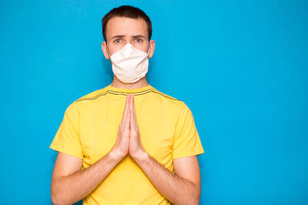 Portrait of young man in yellow t-shirt wearing protective mask against coronavirus, doing pray gesture for health against corona virus is quickly overcome isolated on blue background. Please stay homeの写真素材