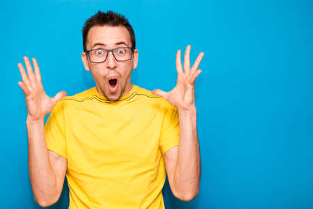 Emotional portrait of man in glasses and yellow t-shirt with shocked, amazed expression isolated on blue backgroundの写真素材