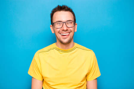 Hfppe portrait of handsome young man laughing in glasses and yellow t-shirt isolated on blue backgroundの写真素材
