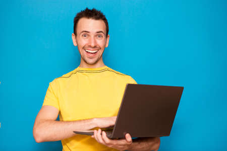 Portrait of excellent smiling young man standing with laptop against blue backgroundの写真素材