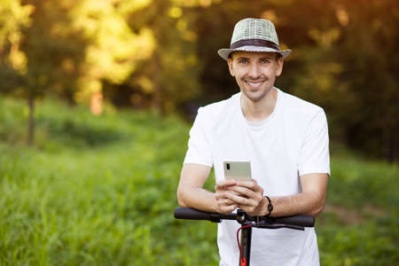 Modern smiling man in stylish white clothes using smartphone while standing at street with electric scooterの写真素材