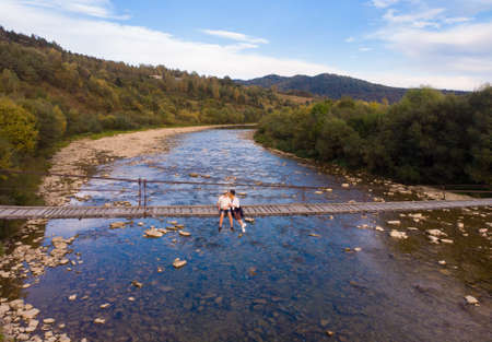 Aerial top view of young love couple sitting on wooden bridge across mountains riverの写真素材