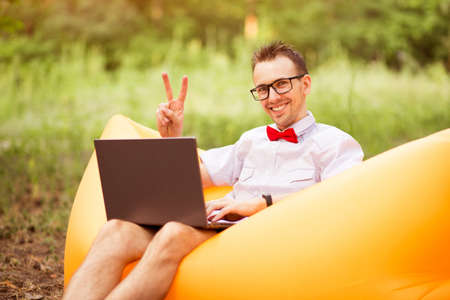 Portrait of handsome successful positive bearded young freelancer in shirt, red bow tie and glasses show v gesture and sitting on inflatable sofa with laptop in summer park. Concept freelancing, teleworkの写真素材