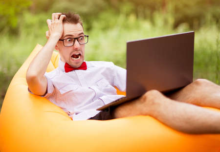 Young freelancer man with shocked face in shirt, red bow tie and glasses sitting on inflatable sofa with laptop in summer park .. Concept freelancing, teleworkの写真素材