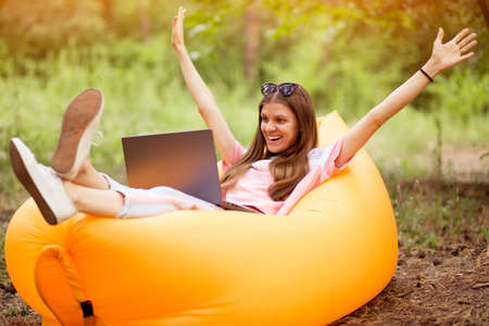 Happy freelancer woman with hands up in winner pose and happy screaming in glasses lying on inflatable sofa with laptop in autumn park .. Teleworkの写真素材
