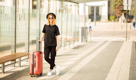 Full length portrait of stylish traveling young woman wear in total black with red suitcase wait for transportの写真素材