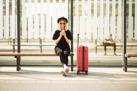Passenger traveler woman with red travel suitcase on stop waiting for air travel, using phone to buy tickets onlineの写真素材