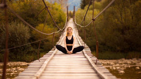Breathing practice. Young fit woman is sitting in yoga butterfly pose on bridge over mountain river. Female is doing yoga stretching exercise or meditation outdoorの写真素材